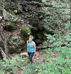 Andrea stands in front of a rocky wall in a forest with small shrubs in the foreground.