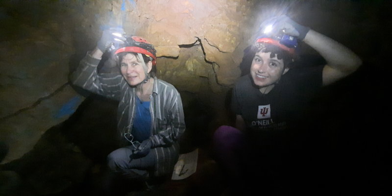 Carla and Gabby look at the camera with caving helmets and flashlights from inside a cave.
